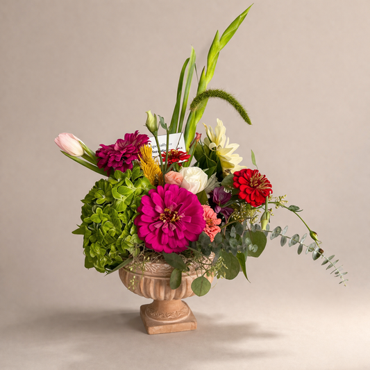 Colorful Mother’s Day floral arrangement in an ornate urn vase featuring zinnias, hydrangea, tulips, roses, eucalyptus, and mixed greenery on a neutral background.